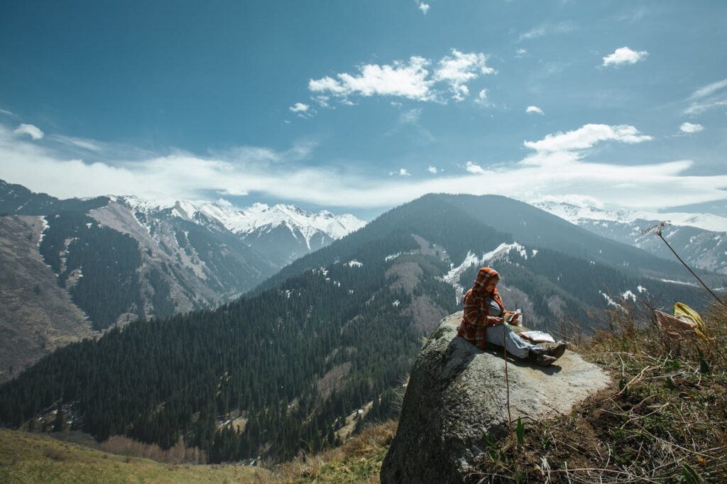 A woman sits on a rock, admiring a breathtaking winter mountain landscape under a clear blue sky.