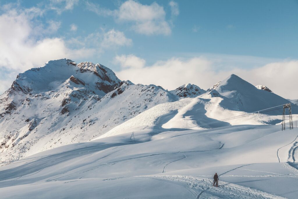 A lone hiker traverses a snowy mountain landscape under clear skies.
